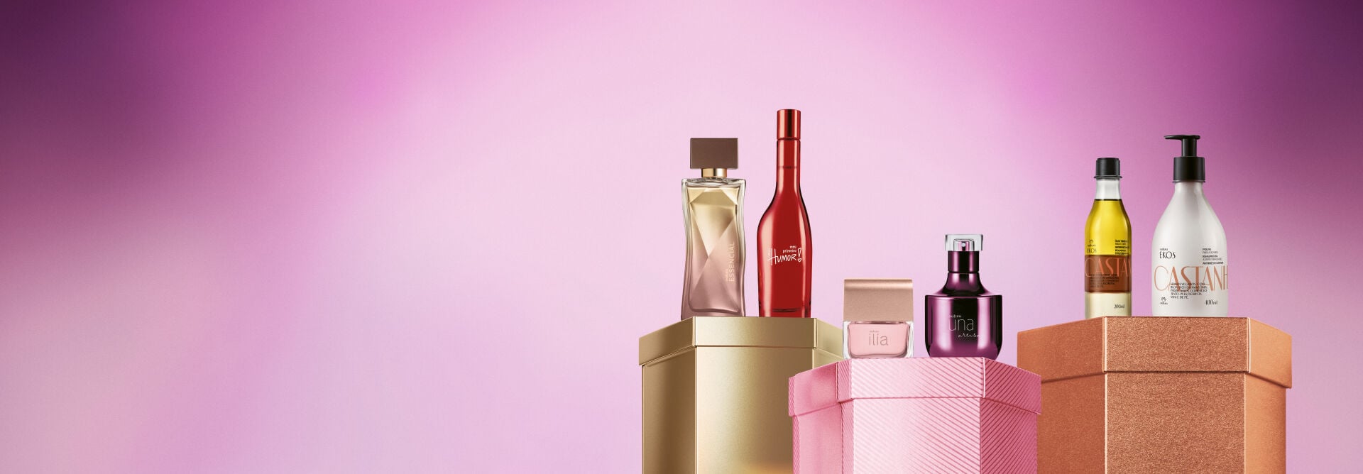 Embalagens Natura sobre caixas brilhantes dourada, rosa e laranja, em fundo rosa: frascos das fragrâncias Essencial, Meu Primeiro Humor, Ilía, Una Artisan e de óleo e polpa para o corpo Ekos Castanha.