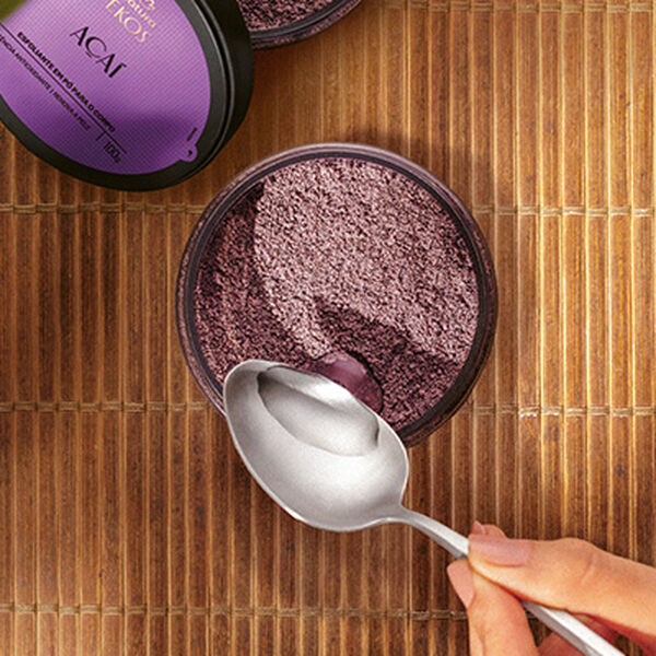 Esfoliante em Pó Ekos Açaí, com embalagem acrílica transparente, tampa preta e rótulo roxo, destampado sobre esteira de madeira. Com uma colher, uma mulher adiciona água ao conteúdo do produto.