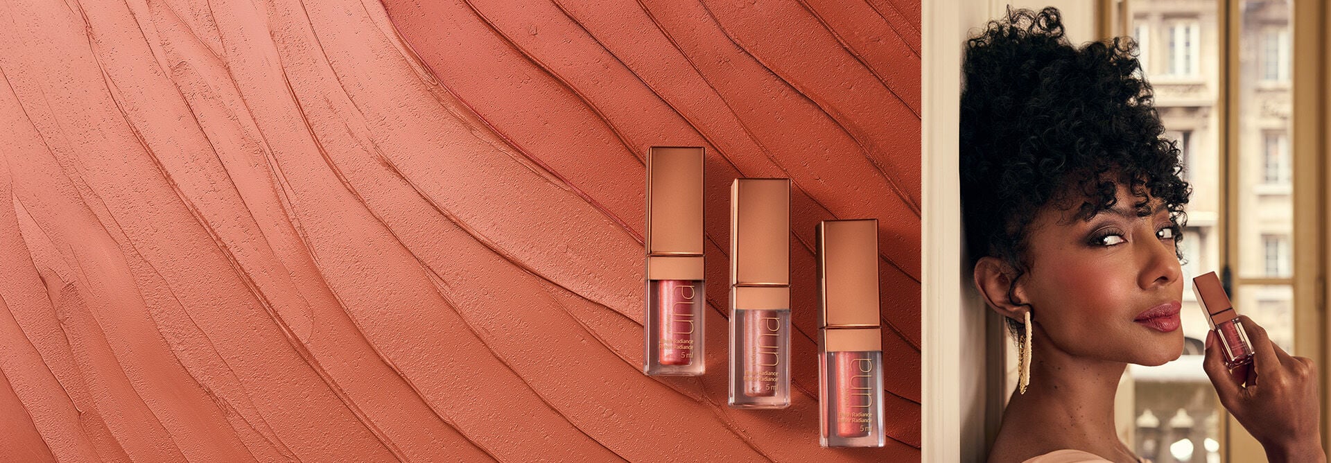 3 embalagens transparentes com tampas nude metalizadas do Blush Líquido Radiance Una, em fundo com produto cremoso rosado. Ao lado, mulher negra de cabelo cacheado segura um frasco do produto.