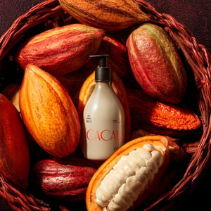  Frasco bege com pump preto de Polpa Desodorante Hidratante Corporal Ekos Cacau 400 ml está entre frutos de cacau inteiros e abertos, dentro de um cesto marrom em fundo escuro.