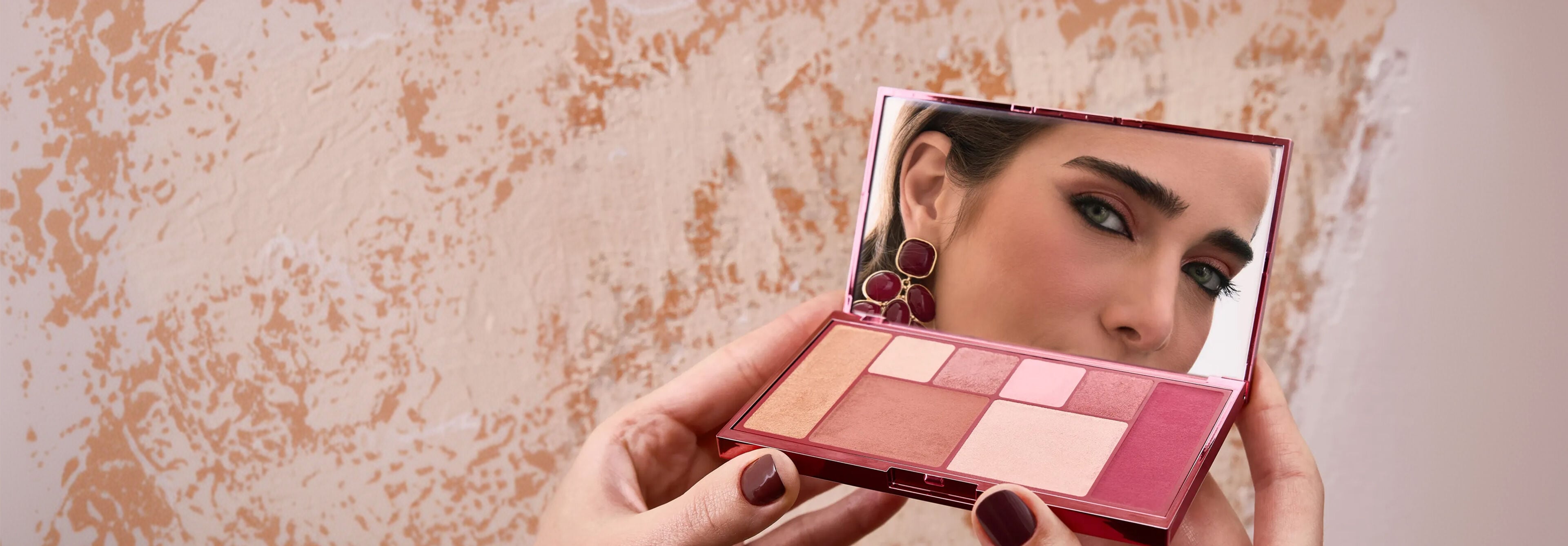Mãos femininas seguram a paleta de sombras aberta da linha Natura Una, refletindo o rosto da modelo clara no espelho interno. O produto destaca tons terrosos e cintilantes.