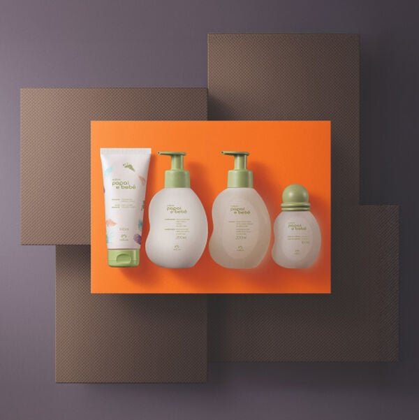 Vista superior de um kit Natura Papai e Bebê centralizado sobre um fundo laranja. Contém uma bisnaga, dois frascos com válvula pump e uma colônia pequena com tampa redonda, todos em tons de verde-claro e bege.
