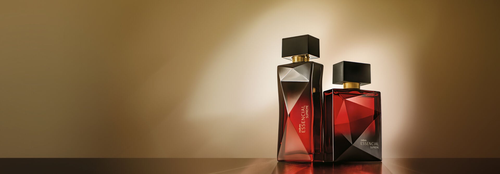 Dois frascos de perfume Natura Essencial Supreme, feminino e masculino, posicionados sobre superfície escura com iluminação focal. Os vidros lapidados em tom vinho transmitem sofisticação.