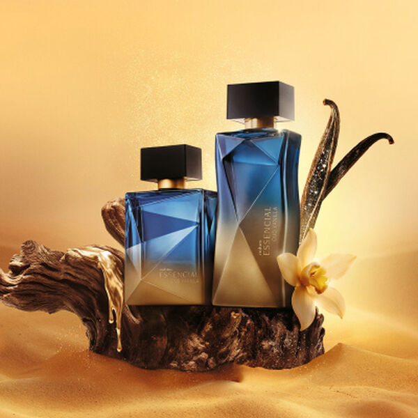 Dois frascos de perfume Natura Essencial Oud Vanilla, um masculino e um feminino, em tons de azul e dourado, sobre uma base de madeira com uma flor de baunilha ao lado.