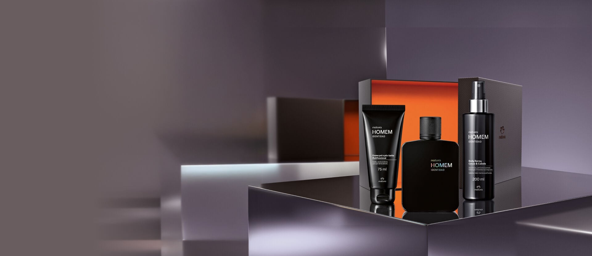 Kit Natura Homem Identidad composto por creme pré e pós-barba, colônia em frasco preto fosco e body spray para corpo e cabelo, posicionados à frente de uma embalagem de presente aberta com fundo laranja.