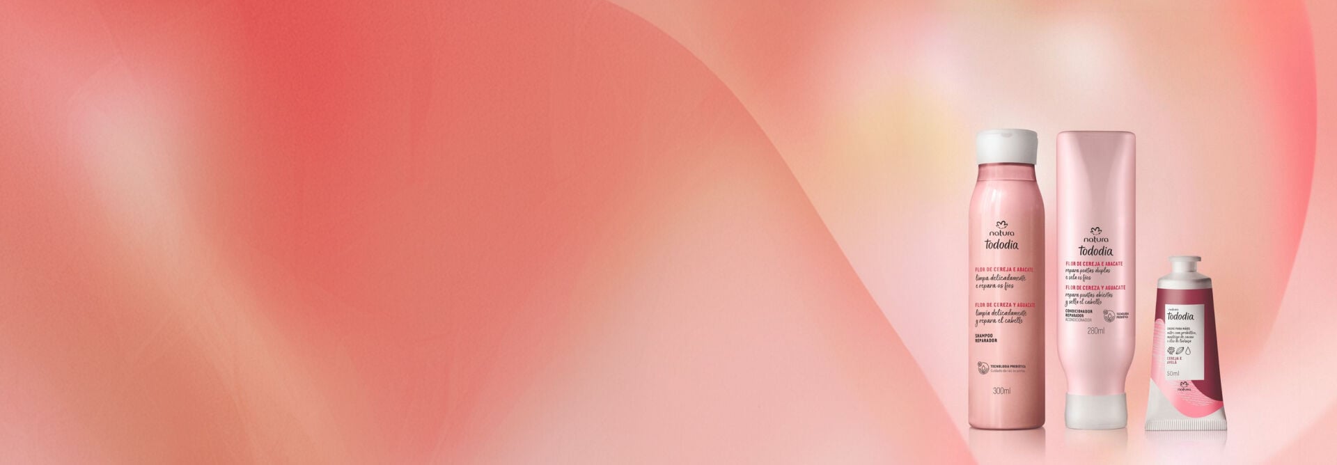 Frascos de Shampoo e Condicionador Tododia Flor de Cereja e Abacate e bisnaga de Creme para as Mãos Tododia Cereja e Avelã, em fundo em tons rosados com elementos gráficos.