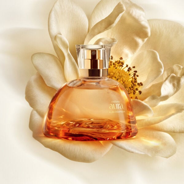 Frasco de perfume Natura Aura Alba em vidro translúcido ambarado repousa no centro de uma grande flor branca aberta. O cenário em tons suaves de creme destaca a elegância da fragrância dourada.