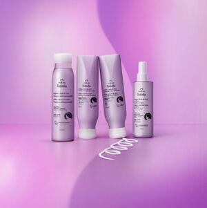 Quatro produtos Natura Tododia Amora e Óleo de Coco para cabelos cacheados e crespos, em embalagens lilás: um shampoo, um condicionador, um creme de pentear e um spray de tratamento. Um rastro de creme em formato de 'S' está na base. Fundo roxo e lilás.