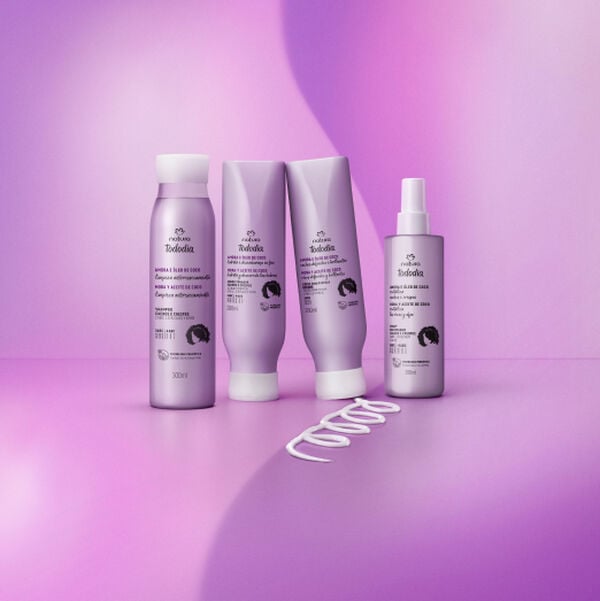Quatro produtos Natura Tododia Amora e Óleo de Coco para cabelos cacheados e crespos, em embalagens lilás: um shampoo, um condicionador, um creme de pentear e um spray de tratamento. Um rastro de creme em formato de 'S' está na base. Fundo roxo e lilás.