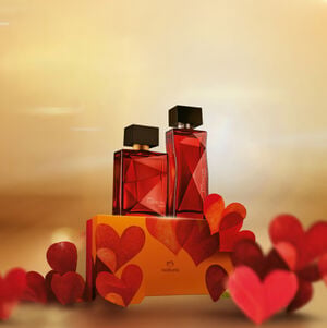 Dois perfumes Natura Essencial Safran em frascos de vidro vermelho lapidado sobre uma caixa de presente decorada. O fundo é dourado e iluminado, com vários corações vermelhos tridimensionais flutuando ao redor, em clima de Dia dos Namorados. 