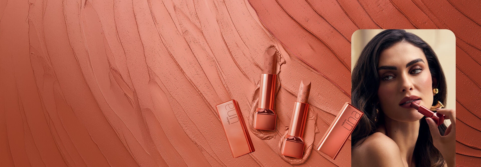 2 embalagens nude metalizadas do Batom CC Hidratante FPS 25 Una abertas, em um fundo com produto cremoso rosado espalhado em ondas. Ao lado, mulher branca de cabelo escuro aplica o batom nos lábios. 