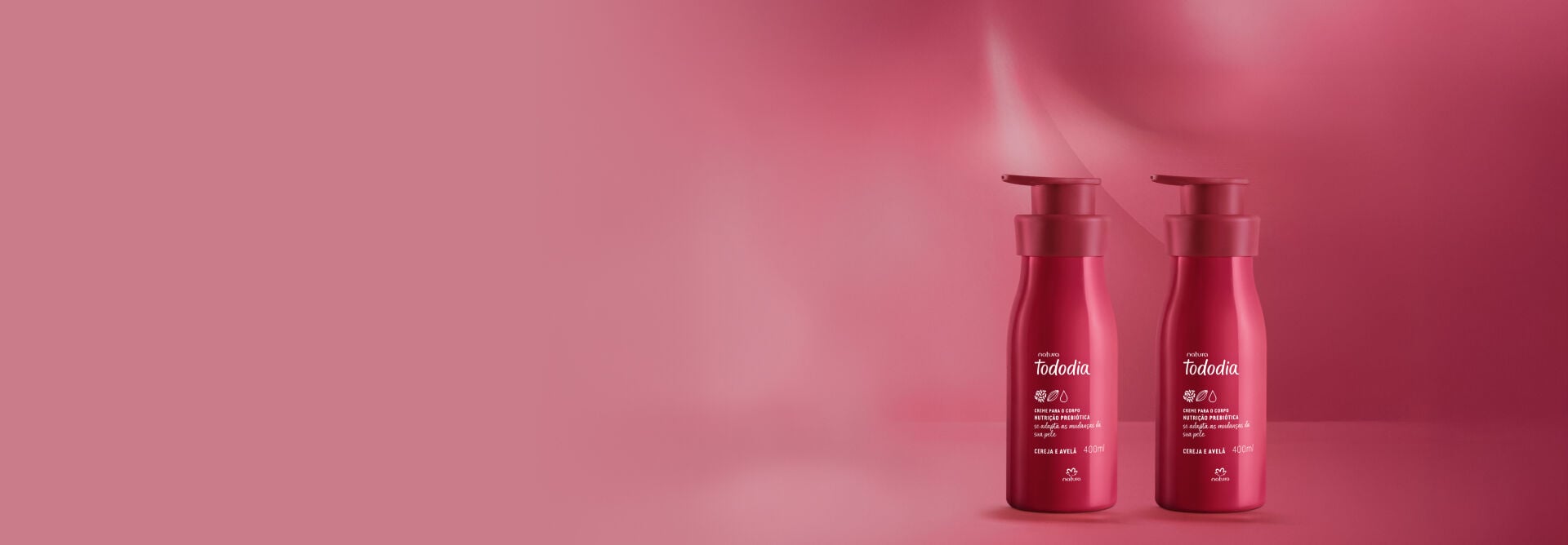 2 unidades do Creme Nutritivo para o Corpo Tododia Cereja e Avelã, com frascos e tampas vermelhos, em fundo rosado.