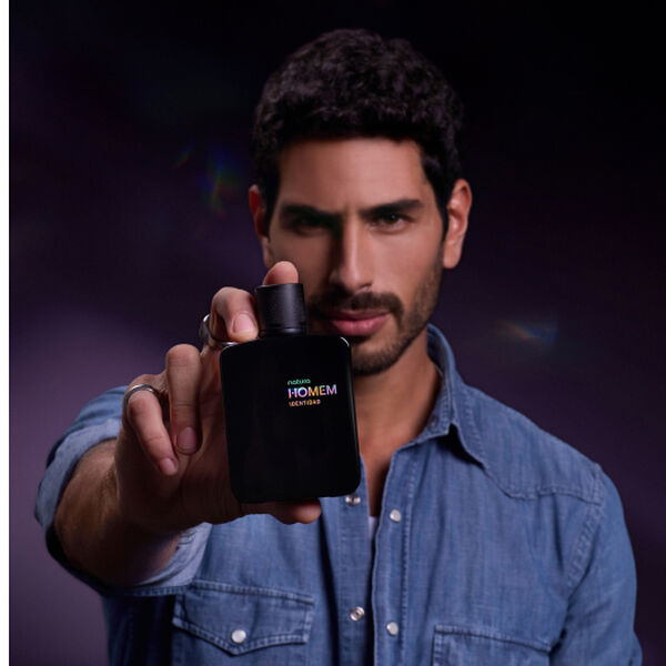 Homem branco, de cabelo escuro e barba bem desenhada, usando camisa jeans azul em fundo roxo-escuro. Ele segura um frasco preto do Deo Parfum Natura Homem Identidad.