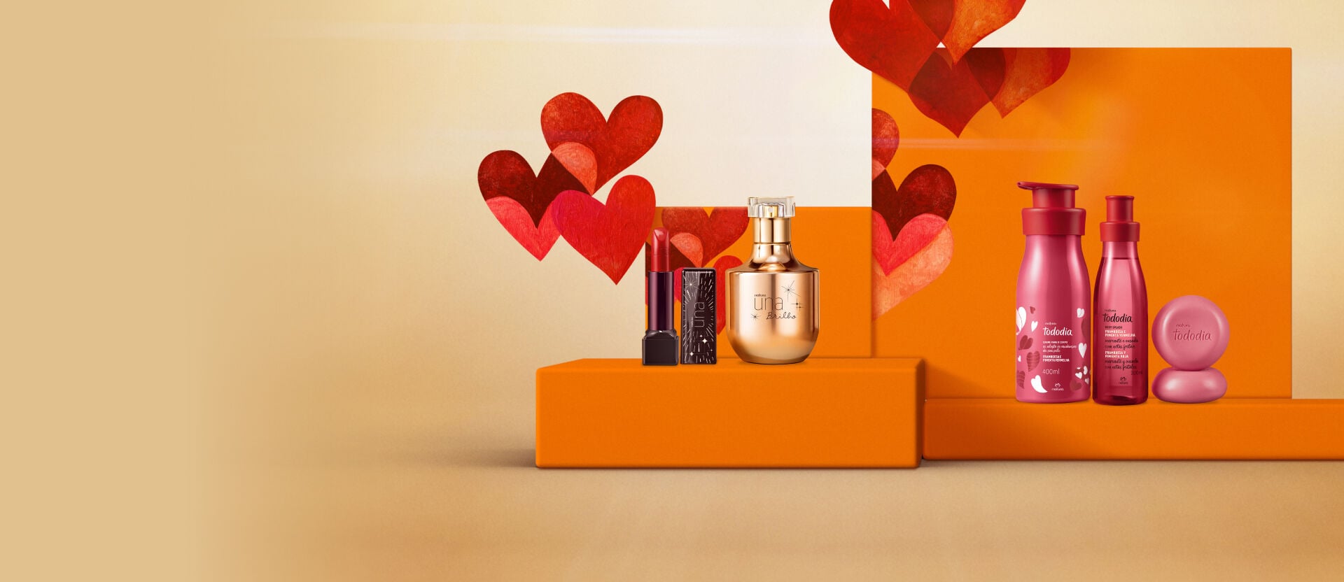 Dois presentes de dia dos namorados Natura em uma bancada laranja com corações ao fundo. À esquerda, um batom e um perfume Natura Una dourado; à direita, três produtos Natura Tododia na cor rosa-escura.