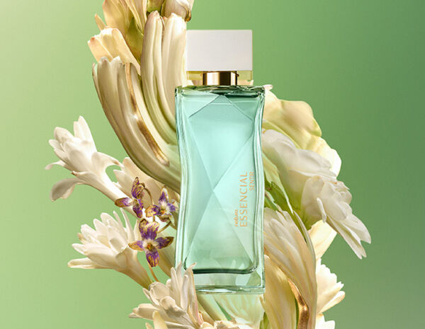 Frasco do perfume Essencial Sentir Feminino, em vidro verde com tampa dourada, posicionado ao centro, cercado por flores brancas e pequenas flores lilases, sobre um fundo em tons de verde degradê.