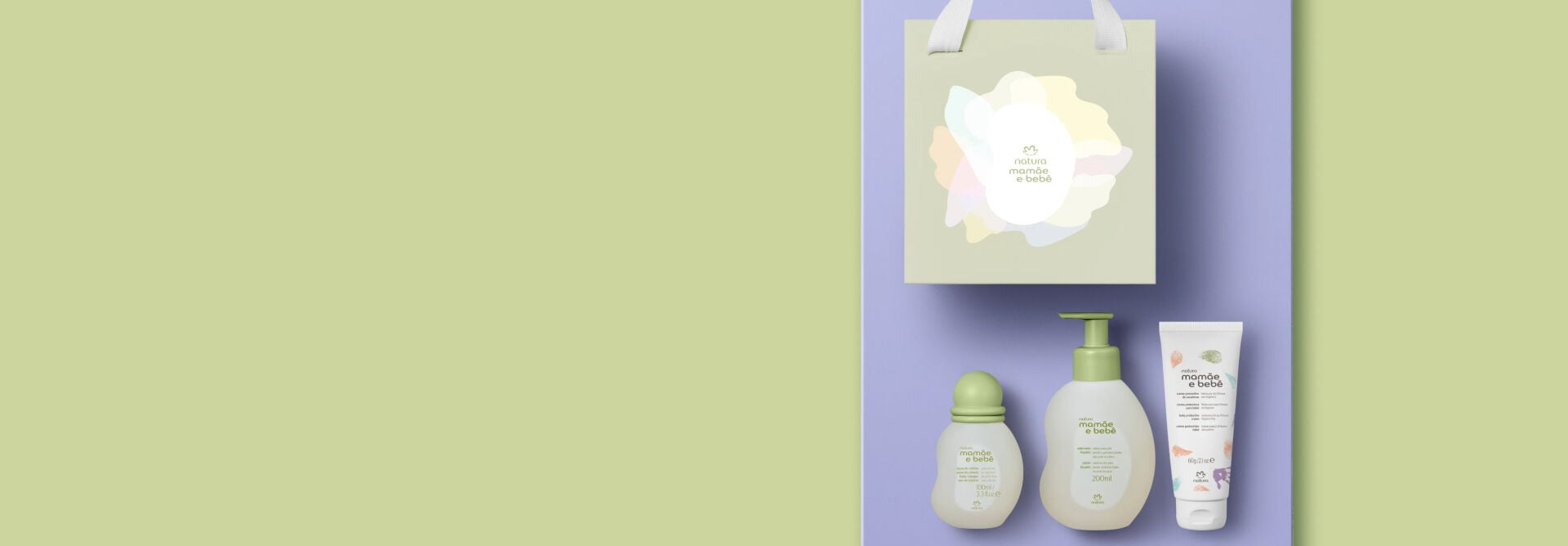 Kit Natura Mamãe e Bebê organizado sobre fundo lilás. Acima, sacola de presente verde-clara; abaixo, colônia, sabonete líquido com pump e bisnaga de creme para assaduras alinhados.
