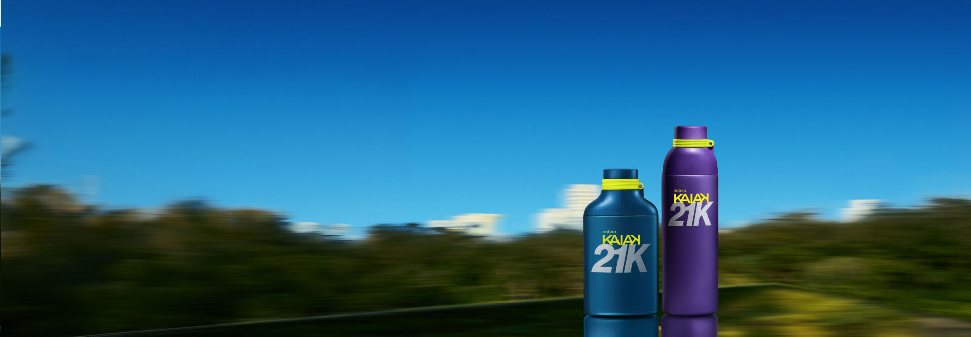 Imagem com fundo em movimento borrado de árvores verdes sob um céu azul limpo. No canto direito, há dois frascos de perfume da marca Natura Kaiak 21K posicionados lado a lado. O frasco da esquerda é mais baixo, largo e possui a cor azul-escuro fosco. O frasco da direita é mais alto, fino e de cor roxa fosca. Ambos têm detalhes em amarelo-limão na tampa e trazem o logotipo "natura KAIAK 21K" .