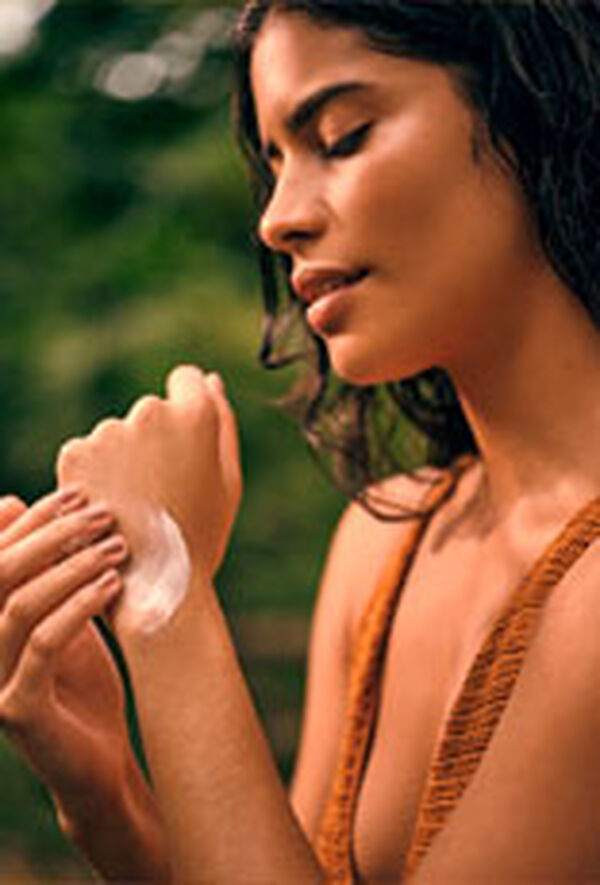 Mulher jovem de perfil aplicando creme branco nas mãos, em um cenário natural com fundo verde desfocado.