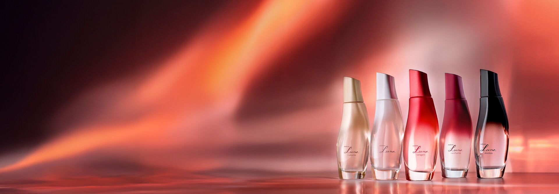 Frascos de fragrâncias da linha Natura Luna, nas cores creme, branco, vermelho, vinho e preto, em fundo em tons de vermelho.