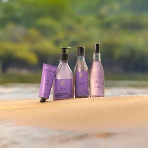 Quatro produtos Natura Ekos Açaí em embalagens lilás estão dispostos sobre a areia, com fundo de rio e vegetação desfocada ao fundo, transmitindo frescor e natureza.