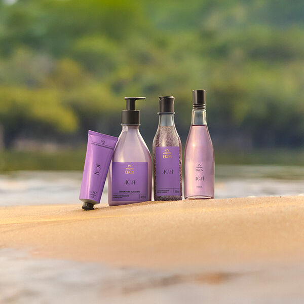 Quatro produtos Natura Ekos Açaí em embalagens lilás estão dispostos sobre a areia, com fundo de rio e vegetação desfocada ao fundo, transmitindo frescor e natureza.