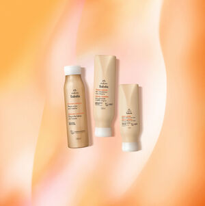 Três produtos Natura Tododia Pêssego e Amêndoa para cabelos. Um shampoo, um condicionador e um creme de pentear em embalagens bege-claro, dispostos em fundo suavemente alaranjado.