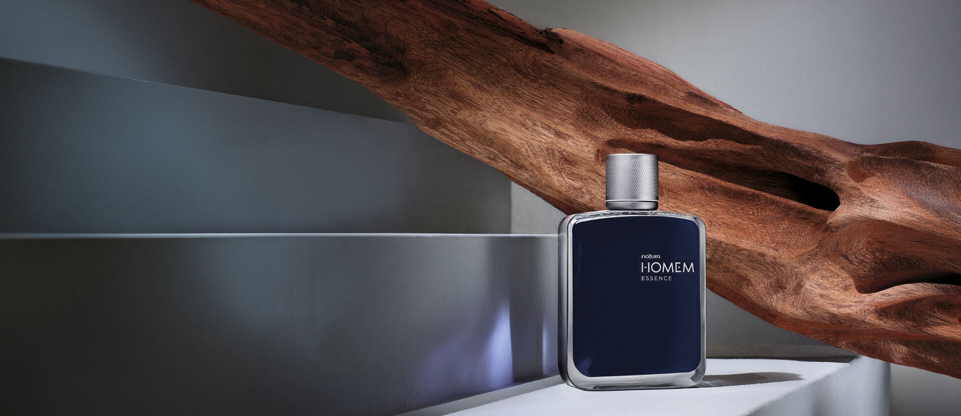 Frasco do perfume Natura Homem Essence sobre base neutra com madeira maciça ao fundo, destacando o design sofisticado da embalagem em azul-escuro e prata.
