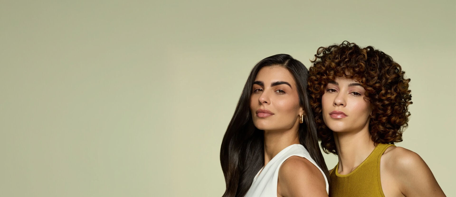 Duas mulheres sobre um fundo verde-oliva claro. À esquerda, mulher de cabelo preto comprido e liso com top branco; à direita, mulher com cabelo castanho volumoso e encaracolado com top mostarda. Ambas exibem maquiagem em tons neutros e suaves.