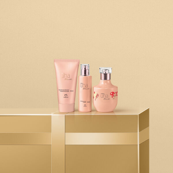 Três produtos Natura Una Blush sob fundo bege sobre caixa de presente dourada. A bisnaga de hidratante, o perfume para cabelos e o frasco de perfume têm embalagens sofisticadas em tom rosa-nude.