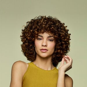 Mulher branca de cabelo castanho cacheado volumoso e brilhante. Ela veste blusa amarela e toca suavemente os fios, em fundo verde-claro.