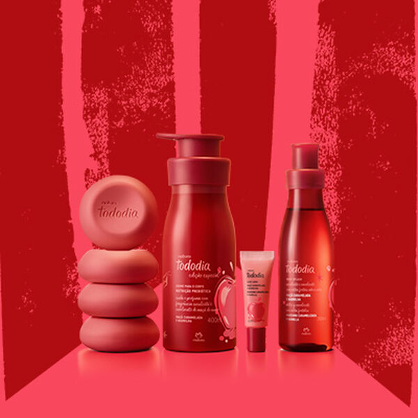 Sabonetes em Barra, Creme Nutritivo para o Corpo, Gloss Labial e Body Splash Tododia Maçã Caramelada e Baunilha, com frascos em tons de vermelho e rosa, sobre fundo vermelho com grafismos cor-de-rosa.