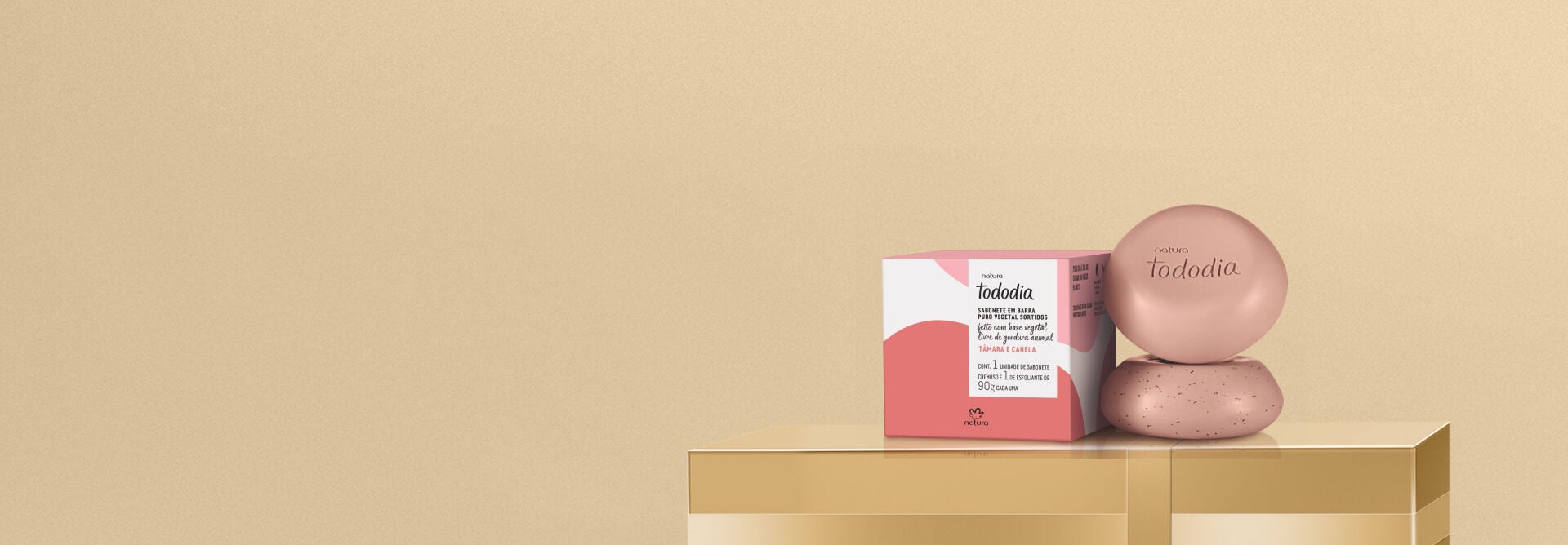 Caixa de sabonetes Natura Tododia Tâmara e Canela sobre caixa presenteável dourada. Ao lado, dois sabonetes arredondados rosa-queimado empilhados, sendo um cremoso e outro esfoliante.