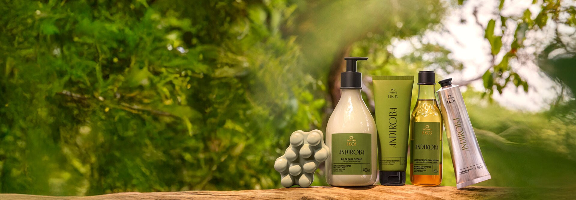 Produtos da linha Natura Ekos Andiroba: Sabonete, Polpa para o Corpo, Hidratante, Óleo e Fluído, sobre madeira, em fundo desfocado com imagem de plantas.