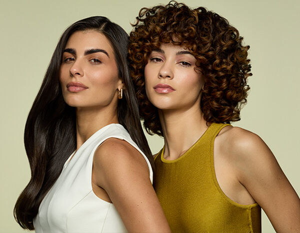 Duas mulheres de pele branca olham para frente, em fundo verde-claro. Uma tem cabelo liso escuro e veste blusa branca, a outra tem cabelo cacheado castanho e veste blusa amarela.