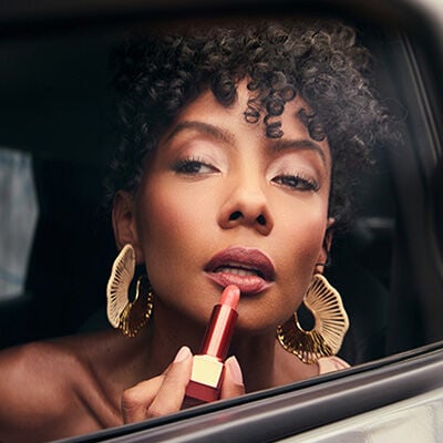 Mulher negra de cabelo cacheado escuro, usando brincos dourados e maquiagem em tons terrosos e rosados, está na janela de um carro. Ela aplica nos lábios um batom com tubo metalizado e bala rosa.