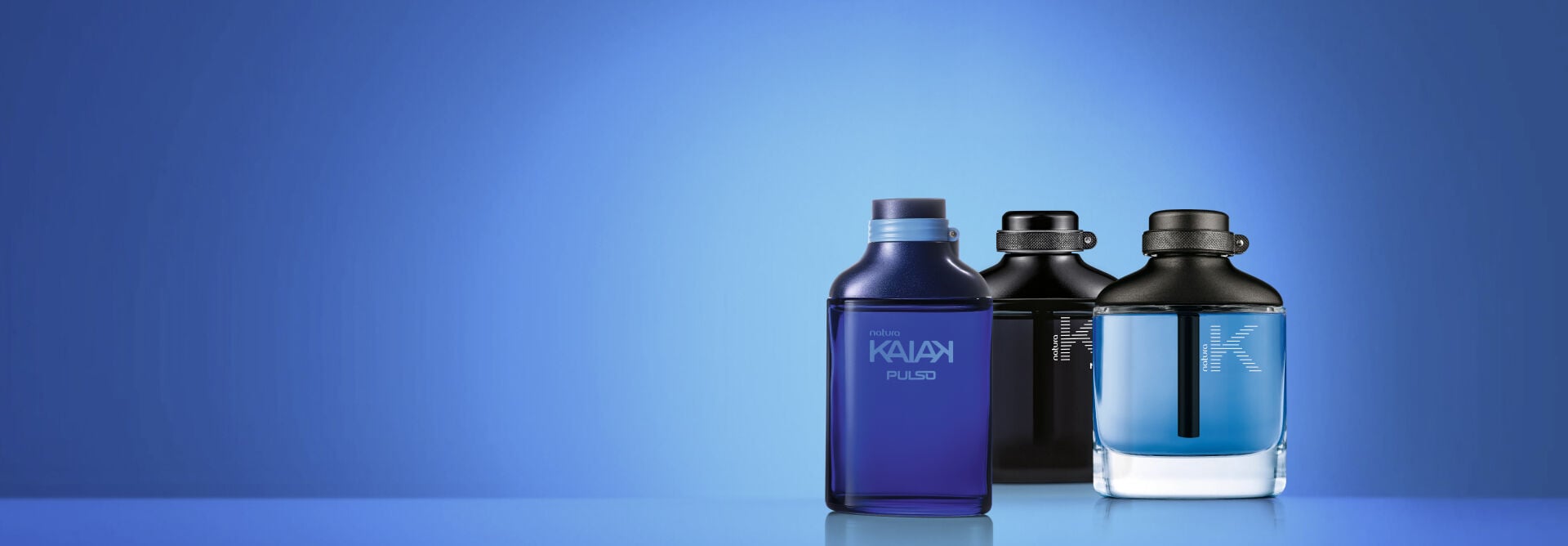 Três Deo Parfums Masculinos da Natura estão lado a lado: Kaiak Pulso à esquerda, Kaiak K Max ao fundo e Kaiak K à direita. Eles estão sobre uma superfície espelhada contra um fundo azul em degradê.
