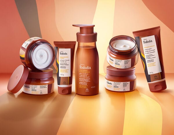 Em fundo em tons de marrom e amarelo, produtos da linha Natura Tododia Jambo Rosa e Flor de Caju: Óleo em Creme, Geleia Desodorante, Loção Corporal, Manteiga Desodorante e Esfoliante Nutritivo.
