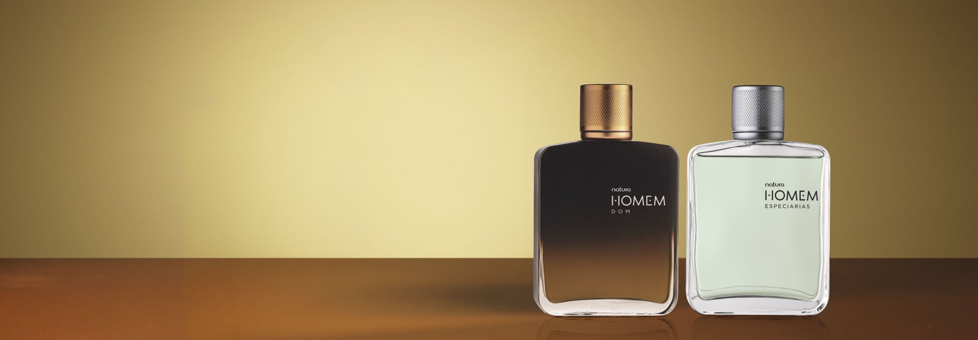 Dois frascos de Natura Homem, Dom e Especiarias, alinhados sobre superfície escura em fundo dourado. As embalagens degradê e translúcida com tampas metálicas refletem elegância e sofisticação.   