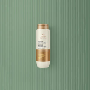 Embalagem creme com tampa e rótulo dourados de Condicionador Antissinais Natura Lumina. Fundo em verde com listras verticais.