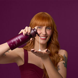 Mulher branca de cabelo ruivo com franja reta sorri em fundo roxo. Ela está apertando a tampa pump do Creme Desodorante Nutritivo Tododia Cereja Negra e Praliné, despejando creme branco sobre a mão.