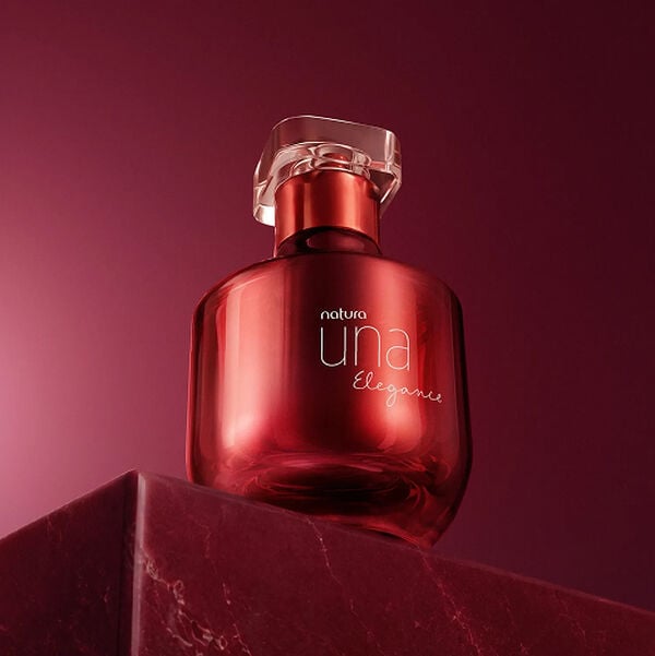 Frasco do perfume Natura Una Elegance em tom vermelho cintilante sobre pódio de pedra em fundo vinho. A embalagem de vidro sofisticada destaca a elegância e a intensidade da fragrância.