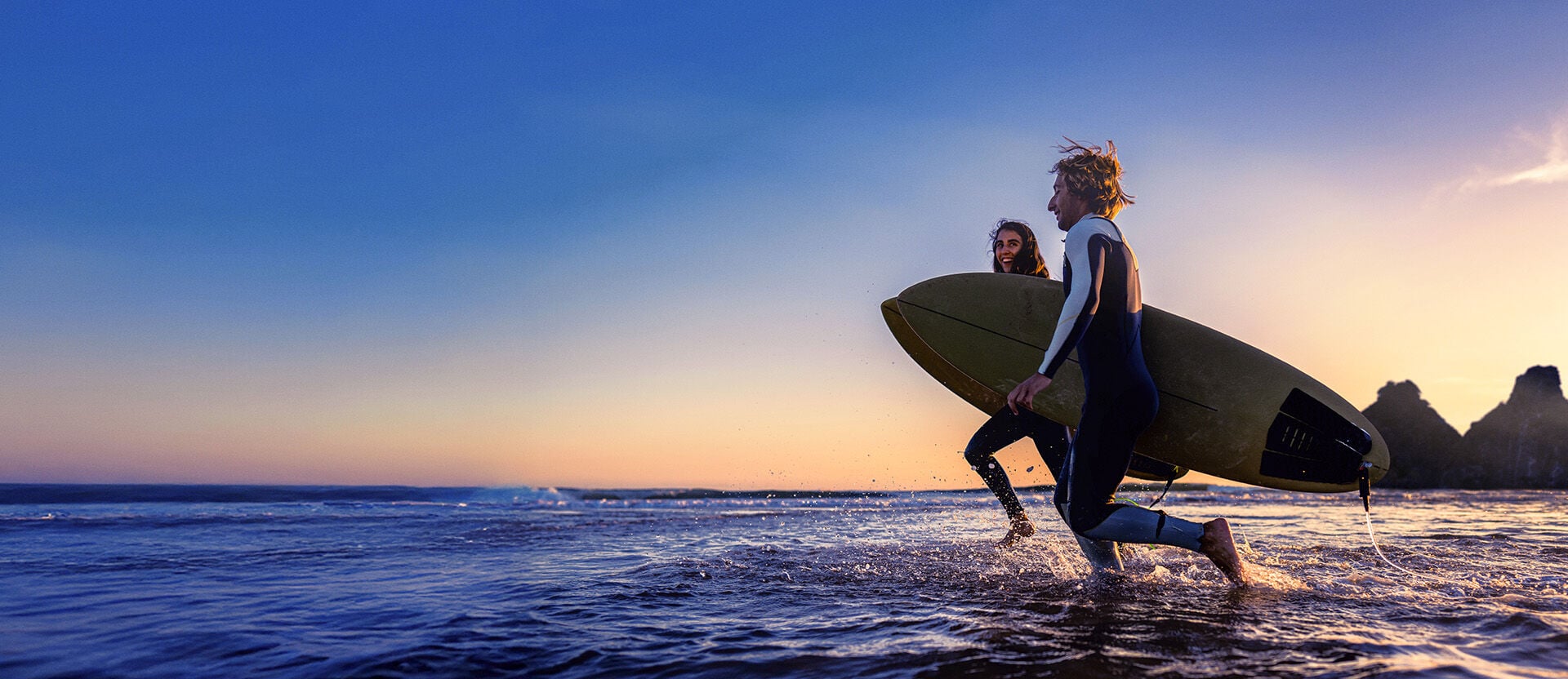 Uma mulher e um homem seguram pranchas de surfe. Eles correm em direção ao mar. Há pôr do sol com céu azul e laranja sobre o mar, com montanhas escuras ao fundo.