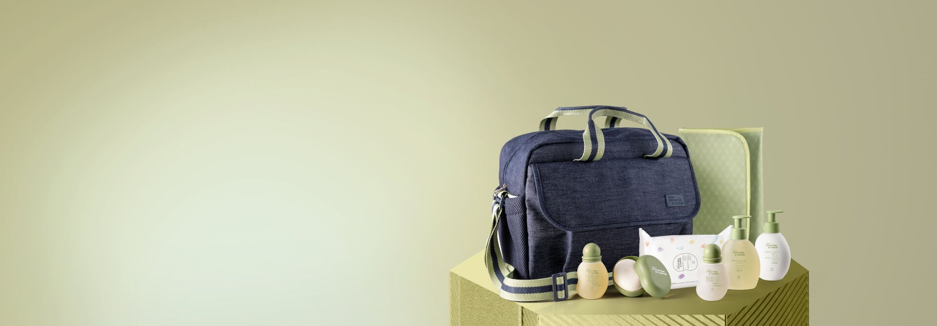Kit Mamãe e Bebê com Trocador sobre caixa verde brilhante, em fundo verde-claro, com: shampoo, condicionador, sabonete com saboneteira, água de colônia, óleo, lenços umedecidos e bolsa com trocador.