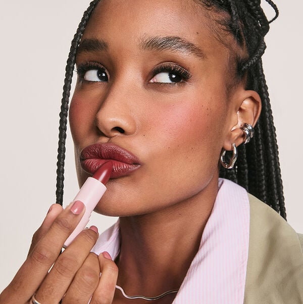 Mulher negra com tranças aplica batom vinho nos lábios, olhando para o lado. Fundo claro.