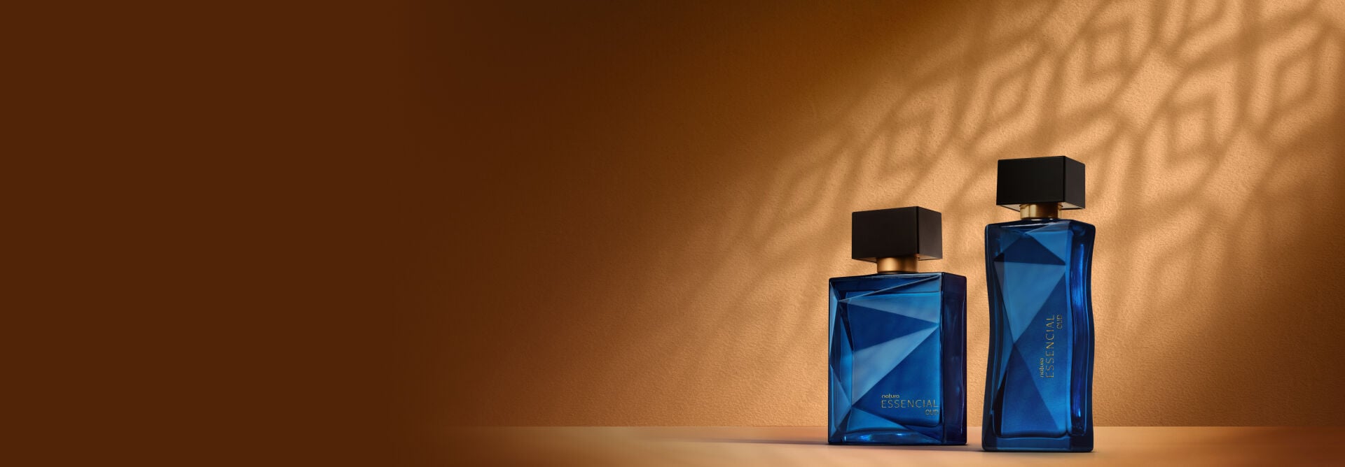 Deo Parfum Essencial Safran Masculino e Deo Parfum Essencial Safran Feminino, ambos em frascos vermelhos com tampas pretas. Estão em fundo cor areia com sombra geométrica projetada.