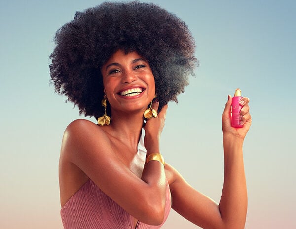 Modelo negra de cabelos crespos usa vestido longo rosê com brincos e bracelete dourados. Ela sorri para a câmera e borrifa o Perfume para Cabelos Luna Divina.