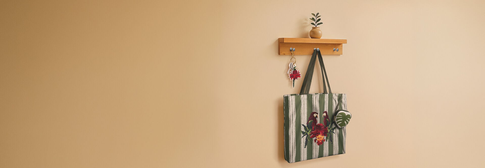Bolsa de tecido com listras verdes e estampa tropical pendurada em cabideiro de madeira na parede beige. O cabideiro também segura chaves com chaveiro de arara e vaso pequeno no topo.