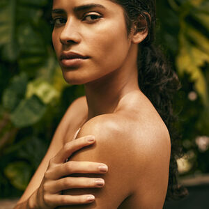 Mulher de pele morena e cabelos castanhos presos olha para a câmera, com uma das mãos apoiada no ombro. Ao fundo, folhas verdes desfocadas sugerem ambiente natural.