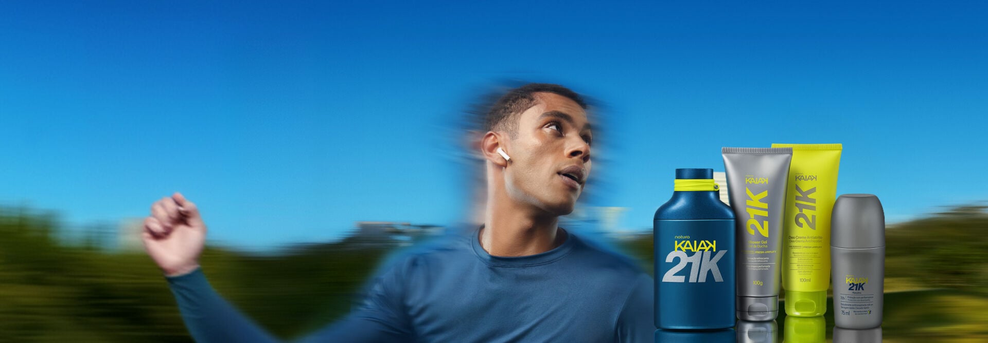 Banner ao ar livre com homem de camiseta azul correndo em velocidade. À direita, perfume azul Kaiak 21K, shower gel cinza, deo creme atrito amarelo e desodorante roll-on cinza.