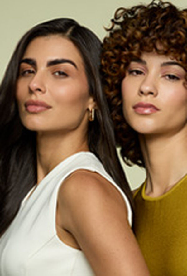 Duas mulheres de pele branca olham para frente, em fundo verde-claro. Uma tem cabelo liso escuro e veste blusa branca, a outra tem cabelo cacheado castanho e veste blusa amarela.